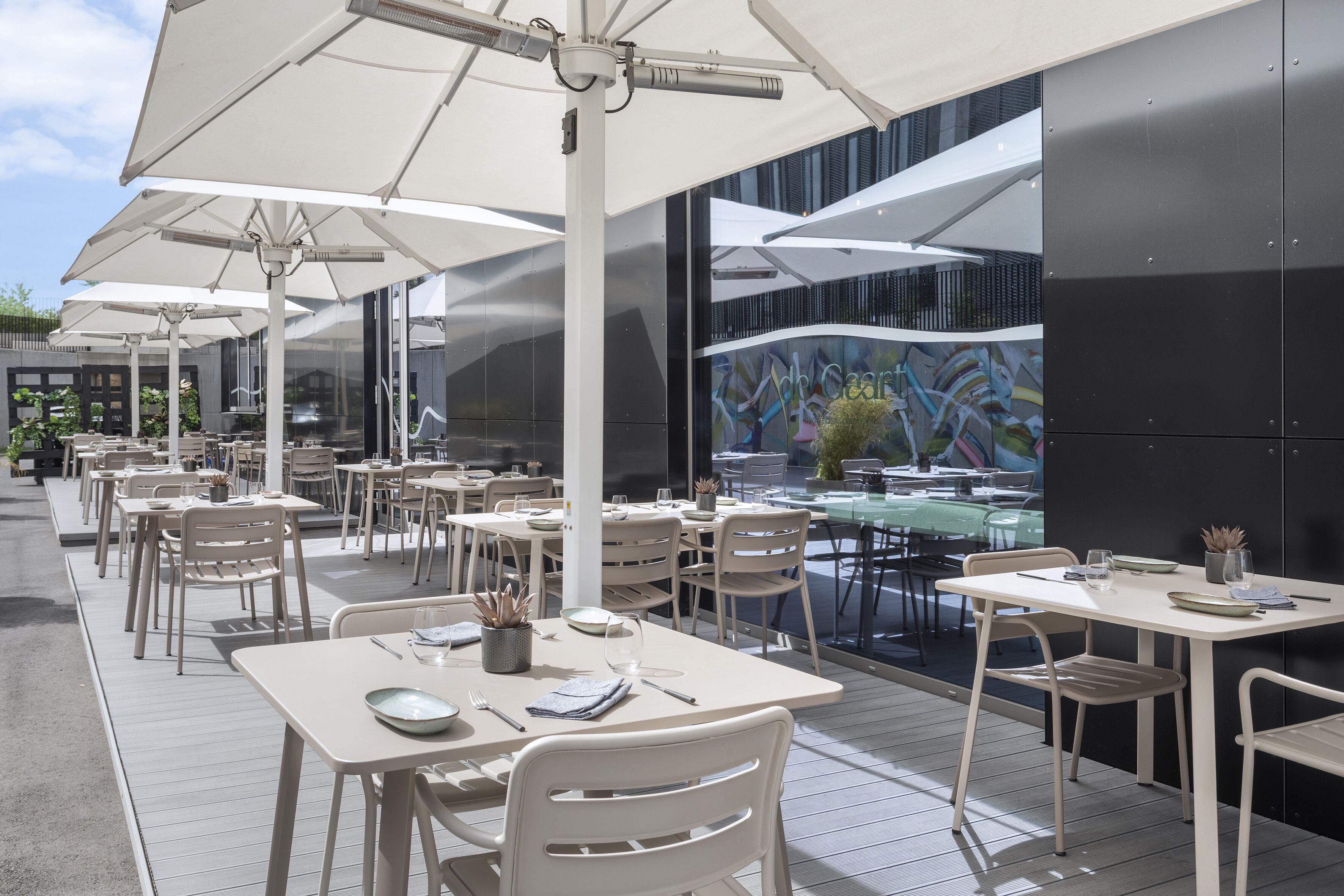 An outdoor dining area with white tables and chairs, large umbrellas, and a modern building with a mural in the background.