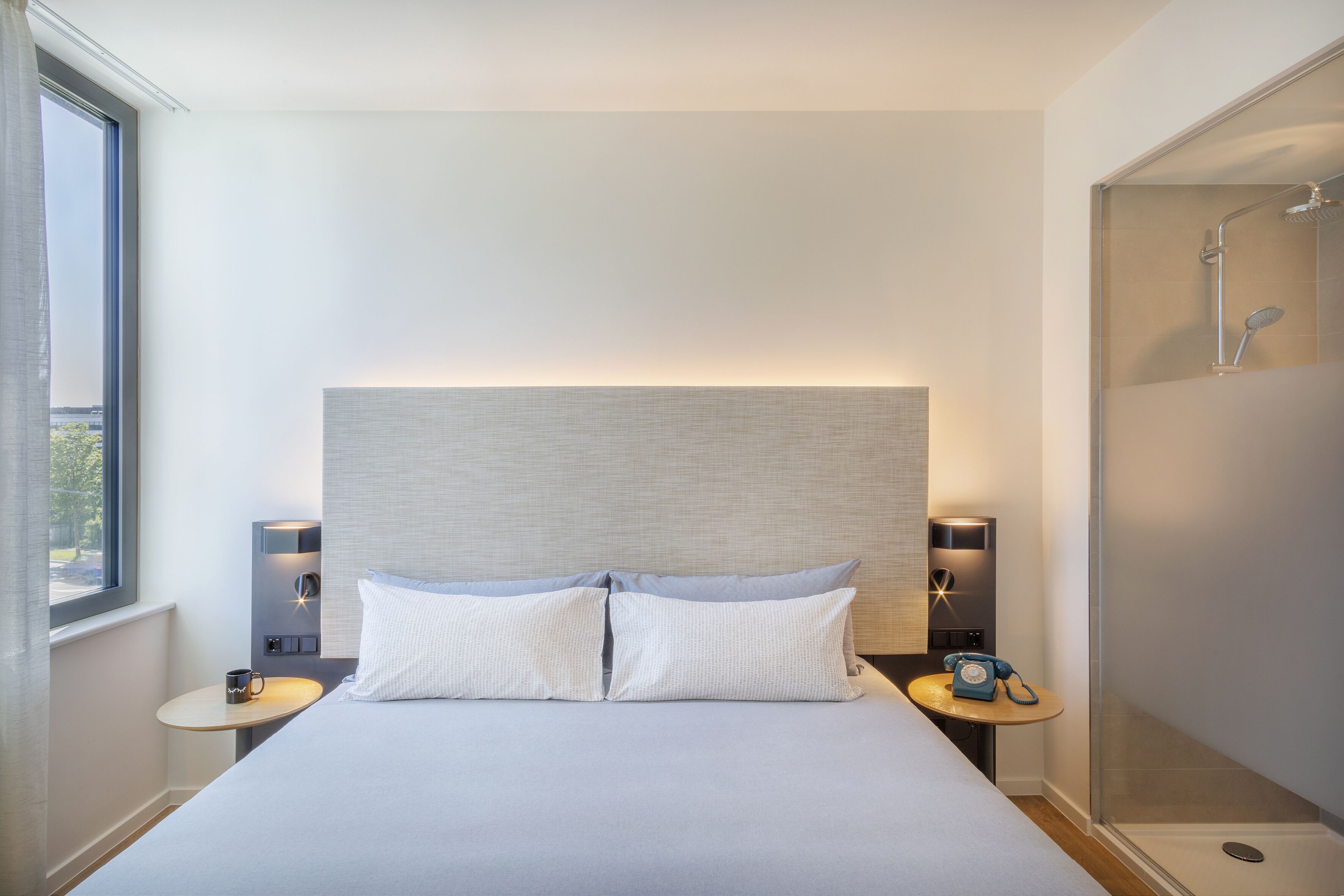 A modern hotel room with a large bed, two bedside tables, a nightstand, a built-in shower, and a window with curtains.