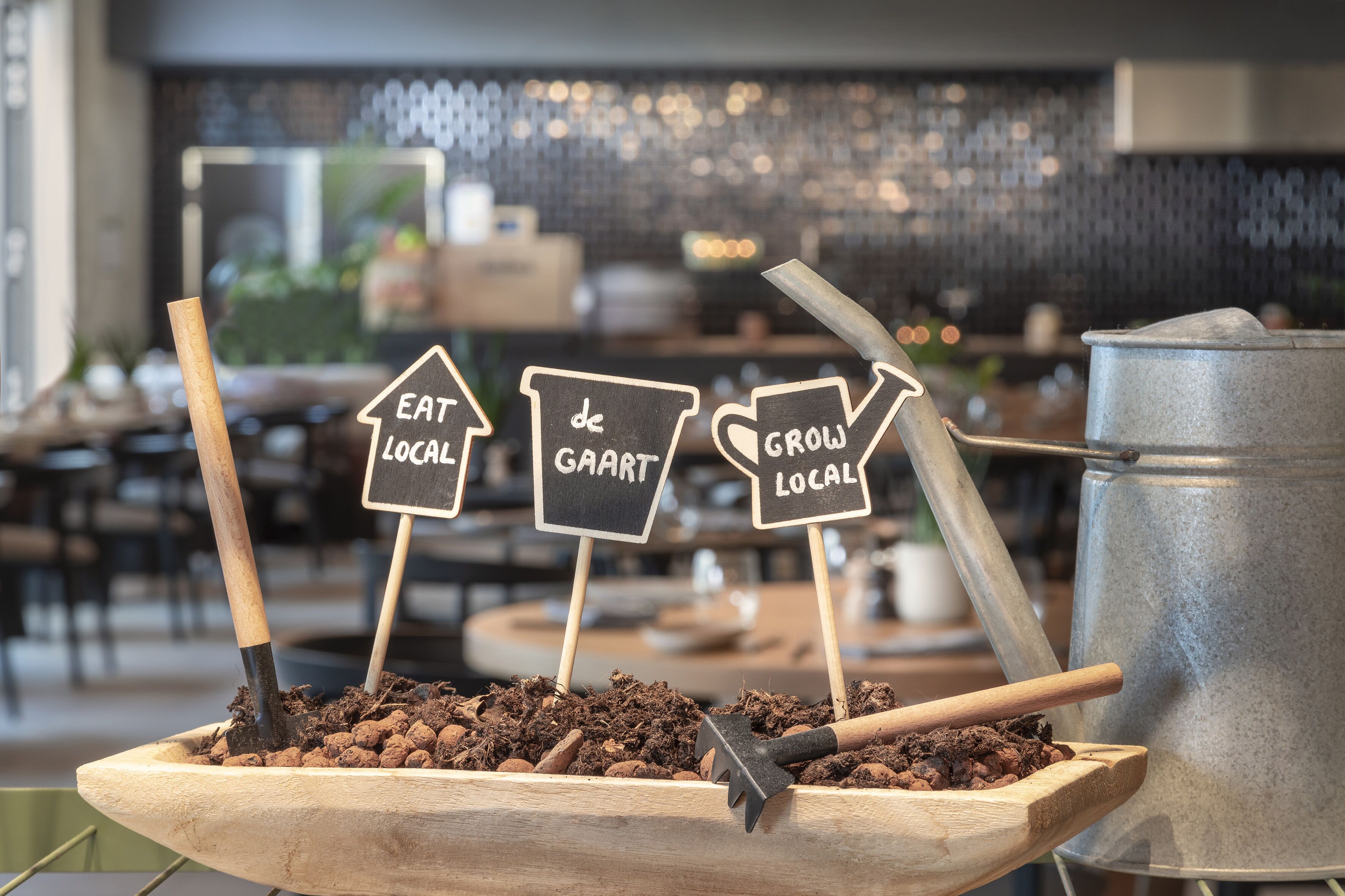 A wooden planter with soil and a shovel, accompanied by signs that read "EAT LOCAL," "de GAART," and "GROW LOCAL."