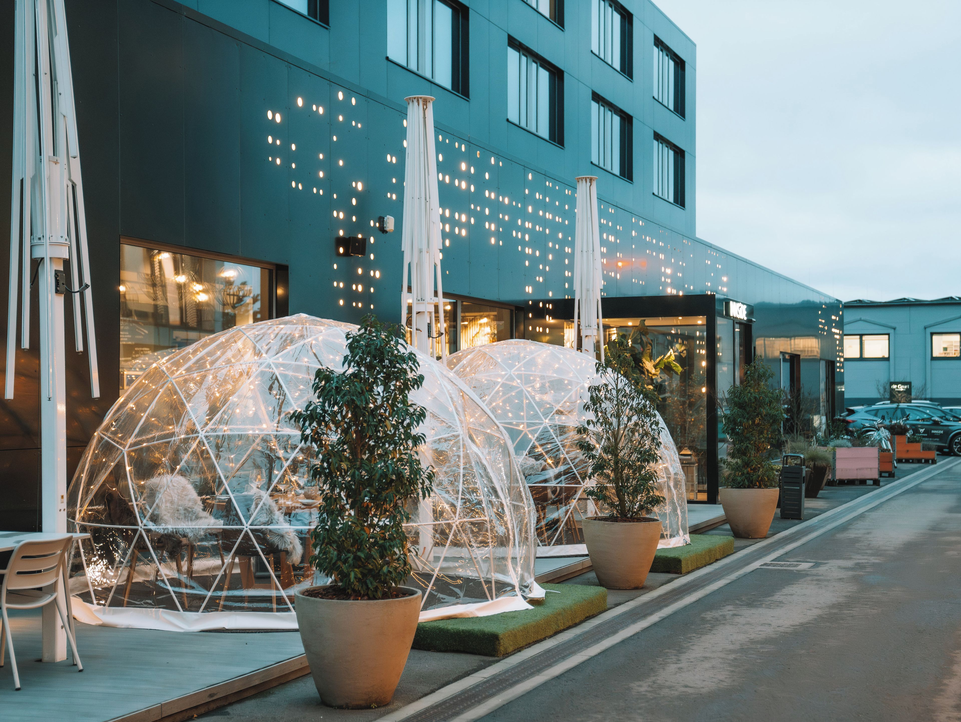 A modern building with large windows, outdoor seating areas with transparent dome structures, and potted plants.