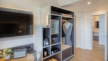 Une chambre d’hôtel moderne dotée d’une télévision à écran plat, d’une armoire intégrée avec des étagères, d’un coin salon et d’une petite table agrémentée d’une plante et d’une radio.