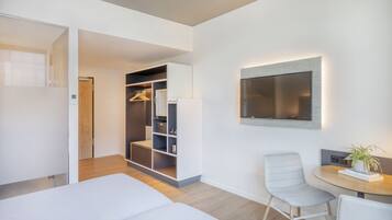 A modern hotel room with a bed, a seating area with a table and chairs, a closet with shelves, and a mounted TV.