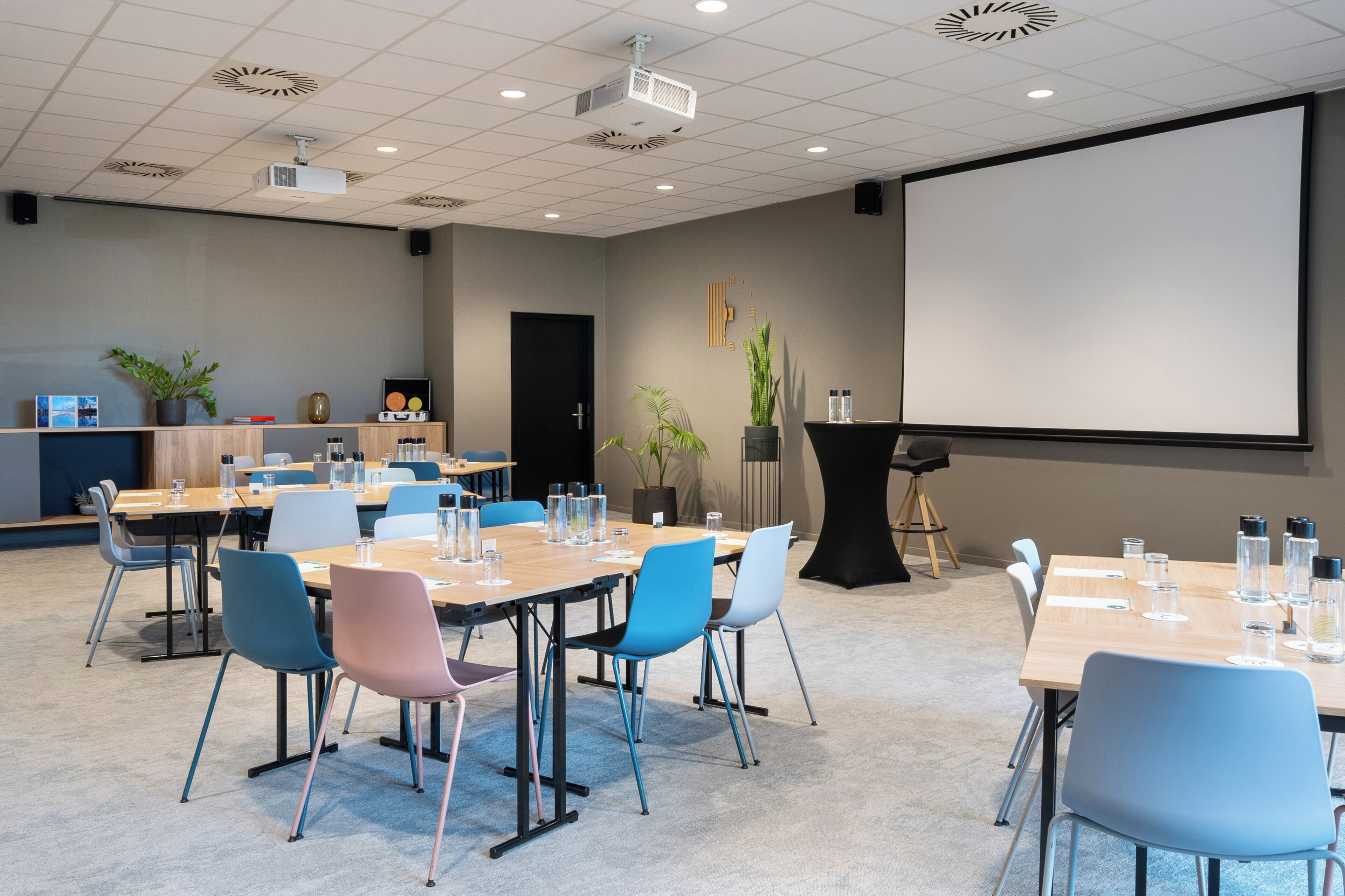 Een vergaderruimte met meerdere tafels en stoelen, een projectiescherm en een presentatiescherm.