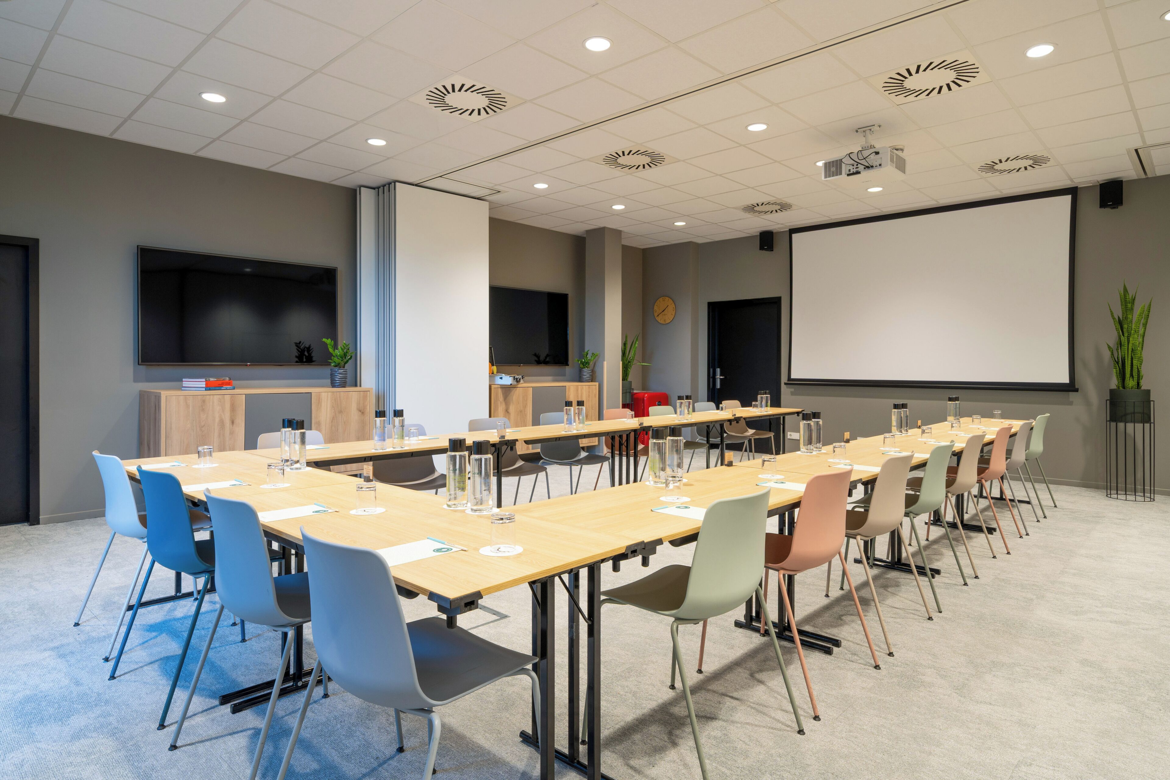 Een vergaderzaal met een lange tafel, stoelen, een projectiescherm en een klok aan de muur.