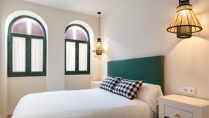 Ein ordentlich bezogenes Bett mit karierten Kissen, ein grüner Kopfteil und ein Nachttisch mit Schubladen. Zwei Fenster mit Vorhängen und eine Wandleuchte sind ebenfalls zu sehen.