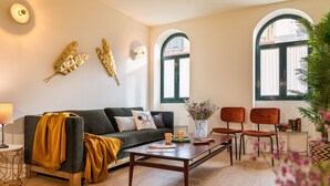Ein modernes Wohnzimmer mit einer Couch, einem Couchtisch und zwei Sesseln. An der Wand befinden sich dekorative Blätter, ein Fenster mit Vorhängen und rechts eine Pflanze.