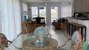 A spacious living room with a glass-top dining table surrounded by wicker chairs, a ceiling fan, and large windows offering a view of the outdoors. - South Beach: A Perfect Beachfront Getaway - Pool, beachfront, ocean views, (Hope Town Bahamas)