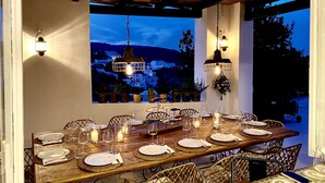 An outdoor dining area with a view of a village at dusk, wooden tables set with plates and glasses, wicker chairs, and hanging lanterns. - Villa "La Quagliara" sea View (Ponza)