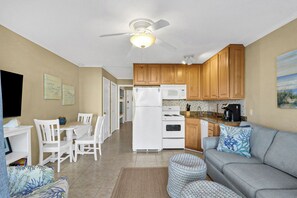 Una cocina compacta con refrigerador, microondas y estufa, un comedor con sillas y mesa, y una sala de estar con sofá y otomanas.