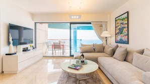 Un salon moderne avec un canapé, une table basse et un téléviseur. Il y a un balcon avec vue sur la mer.