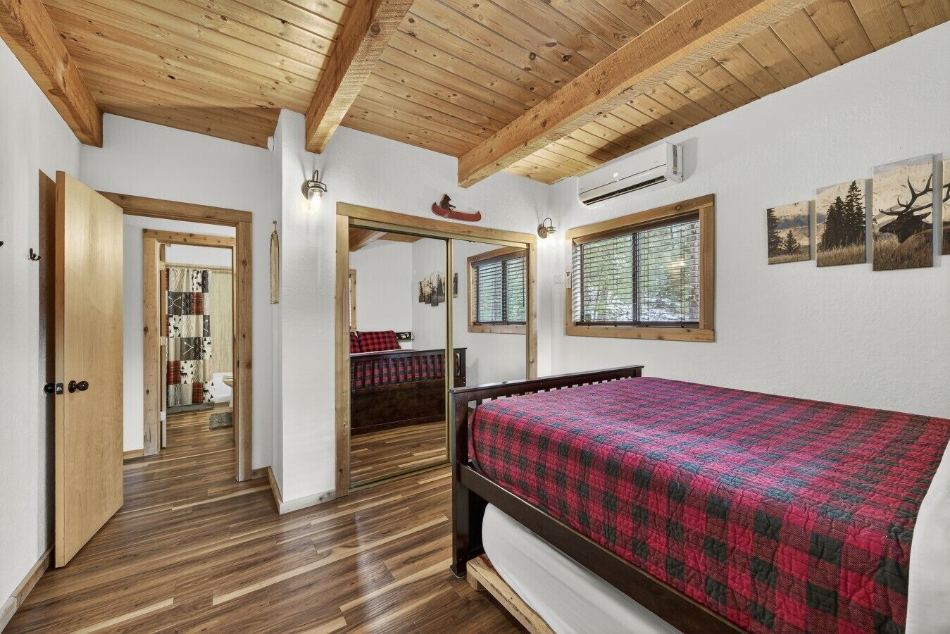 A bedroom with a wooden ceiling, a bed with a red and black checkered bedspread, a window with a view of trees, and an air conditioning unit on the wall.