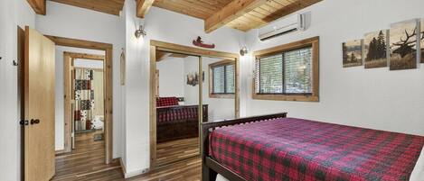 A bedroom with a wooden ceiling, a bed with a red and black checkered bedspread, a window with a view of trees, and an air conditioning unit on the wall.