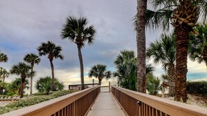 A wooden boardwalk leading to a beach with palm trees and a sign reading "For ecological reasons, the dune."