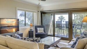 A living room with a ceiling fan, a television displaying a vacation ad, a sofa, a coffee table, and a view of the outdoors through a sliding glass door.
