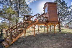 Une cabane en bois perchée dans un arbre, avec un escalier et une petite plateforme, entourée d’arbres.