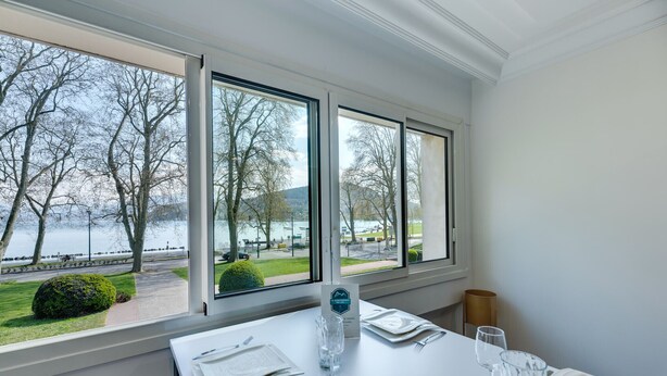 A dining table set with a water bottle, glasses, and cutlery, with a view of a lake and trees through large windows. - Swan Lake - Design apartment facing Lake Annecy (Annecy)