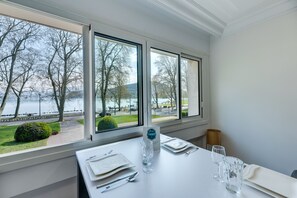 A dining table set with a water bottle, glasses, and cutlery, with a view of a lake and trees through large windows.