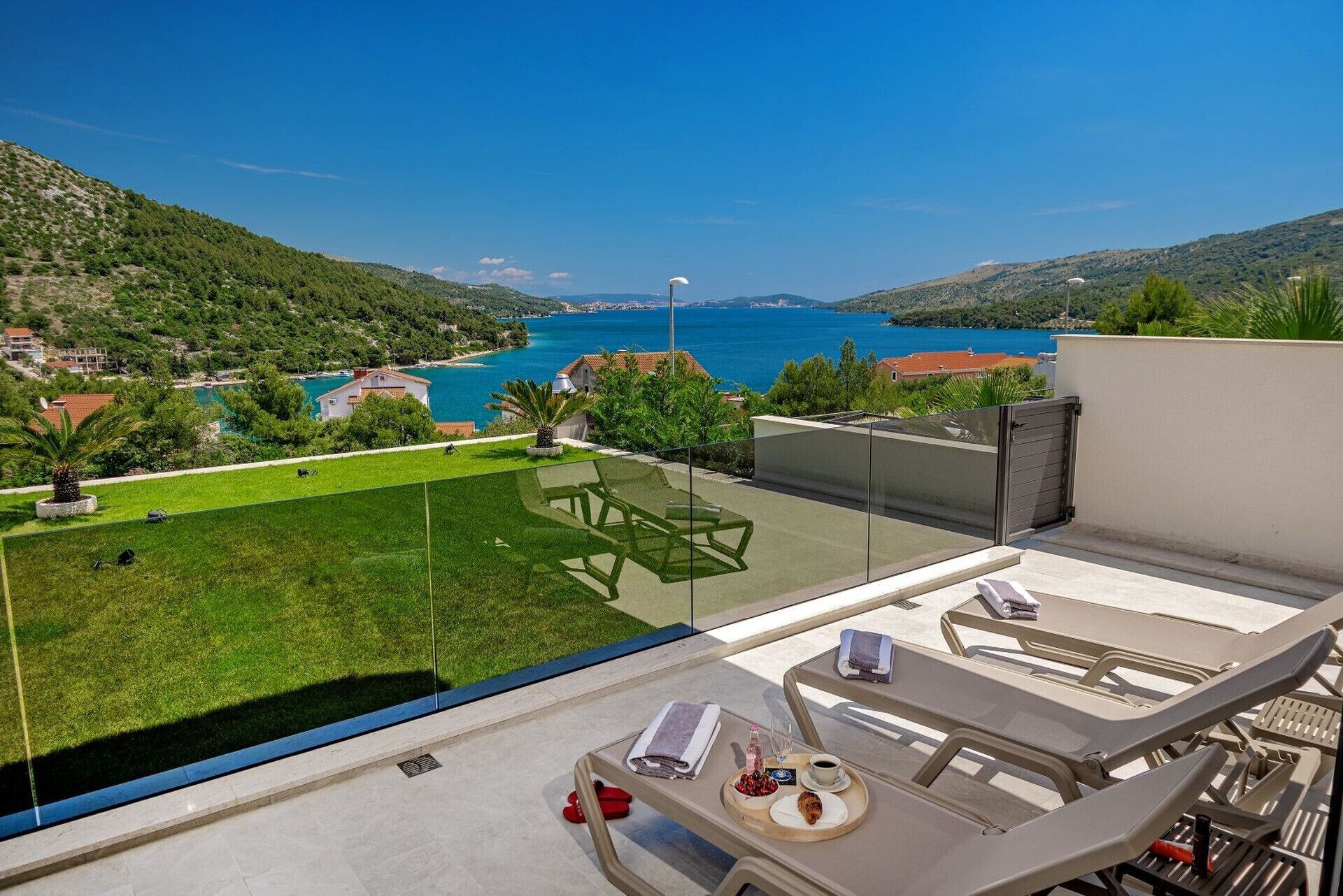 A balcony with a view of a coastal landscape, a dining table with a tray of food, and a chair.