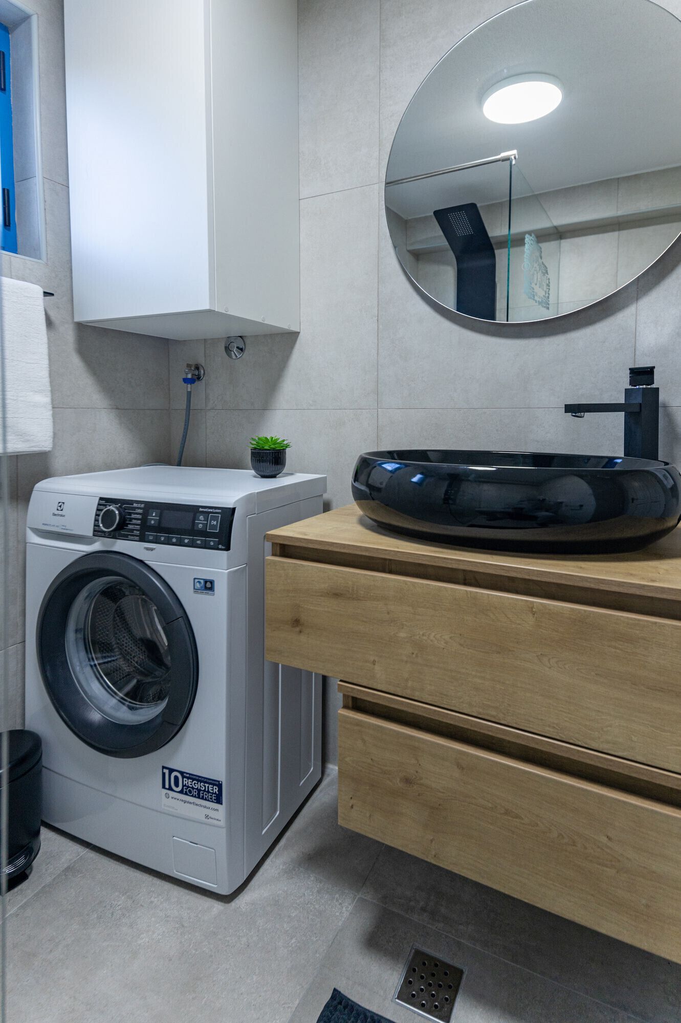 A compact laundry area with a front-loading washing machine, a wooden vanity with a sink, and a round mirror above the vanity.