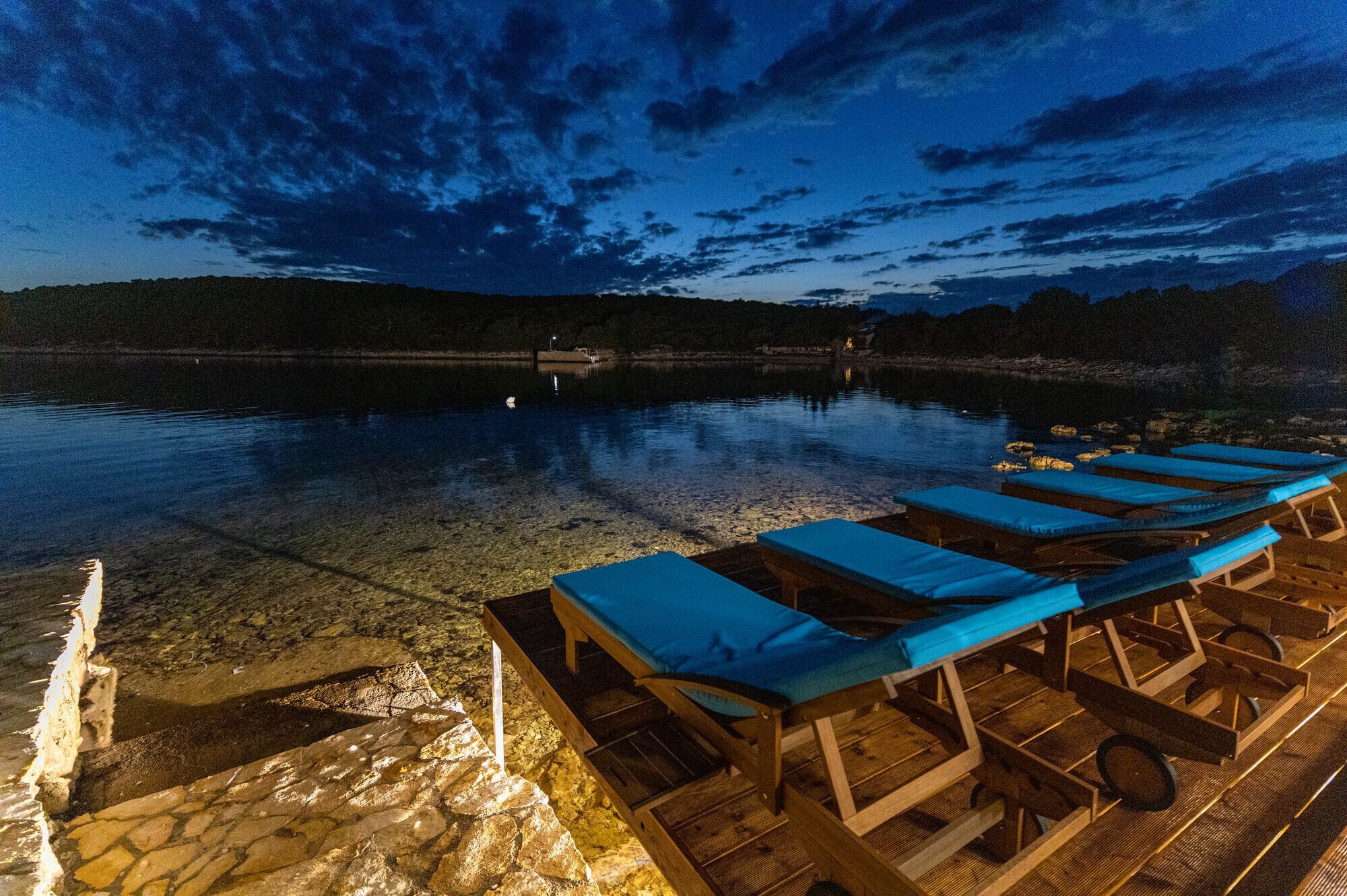 Ein Nachtblick auf eine See-Lounge mit klarem Wasser, einer Holzterrasse und Liegestühlen.