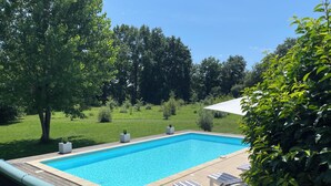 A rectangular swimming pool with a wooden deck and tiled border, surrounded by lush greenery and trees.