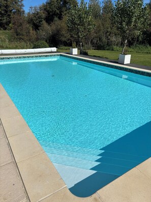 Una piscina rectangular con agua azul cristalina, rodeada por una terraza con baldosas y una zona verde con árboles.