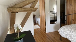 A bedroom with wooden beams, a bed with a floral duvet, a small table with a potted orchid, and a mirror reflecting a room with a bed and a lamp.