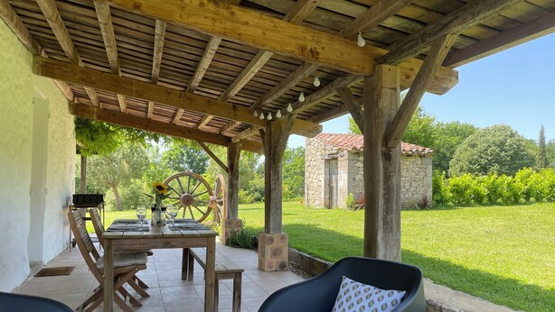A covered outdoor patio with a wooden table set for dining, a stone building in the background, and a grassy lawn.