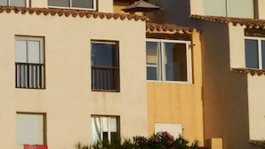 Un edificio de varios pisos con techo de tejas, múltiples ventanas y un pequeño jardín con flores rosadas.