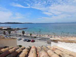 A coastal view with a beach, parked cars, and a road leading to the sea. - Logement en Copropriété Avec Terrasse Face à la Plage (Six Fours les plages)