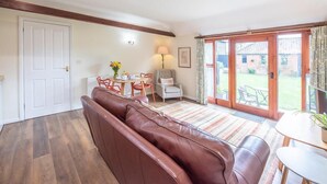 A cozy living room with a brown leather sofa, a dining table with chairs, a lamp, and a view of the backyard through a sliding glass door. - Stables at Valley Farm Barns, Snape (Saxmundham)