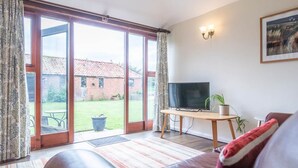 A living room with a TV on a wooden stand, a sofa, a small table with a plant, and a view of a garden through large sliding doors. - Stables at Valley Farm Barns, Snape (Saxmundham)