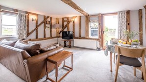 A cozy living room with a leather sofa, wooden beams, a television, and a dining area with a table set for breakfast. - Hayloft at Valley Farm Barns, Snape (Saxmundham)