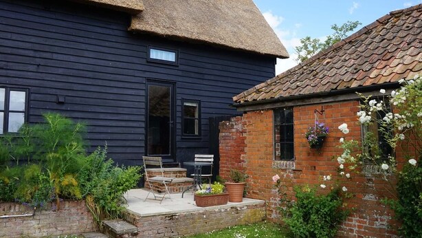 A dark wooden house with a thatched roof and a brick extension. There is a small patio with a table and chairs, and a flower box on the brick wall. - Hayloft at Valley Farm Barns, Snape (Saxmundham)