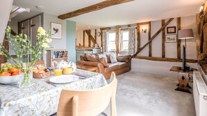 A cozy living room with a high ceiling and exposed wooden beams, a dining area with a table set for breakfast, and a comfortable sofa. - Hayloft at Valley Farm Barns, Snape (Saxmundham)