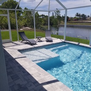 Piscina cubierta con dos sillas de descanso y una mesa pequeña, rodeada por una pérgola blanca y vista a un lago y vegetación.