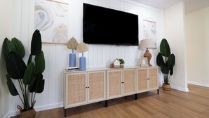 A living room with a flat-screen TV mounted on a white paneled wall, a wicker console table with decorative items, and large green plants.