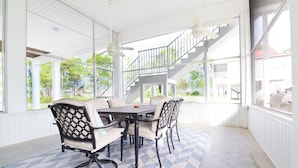 A covered patio area with a dining table and chairs, a staircase, and large windows offering views of greenery.