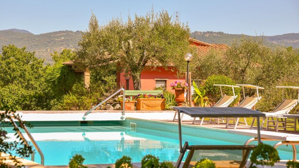 A swimming pool with lounge chairs, surrounded by greenery and a building with a red roof.