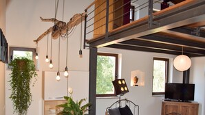 A modern living room with a wooden dining table, a sofa, and a TV. There are hanging lights and a plant on the wall.