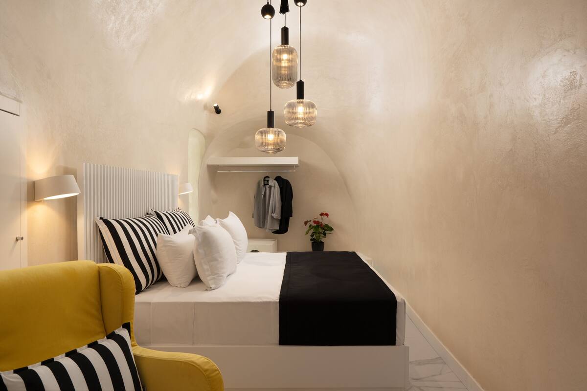 A compact bedroom with a bed, a yellow armchair, a bedside table, a lamp, and a hanging light fixture.