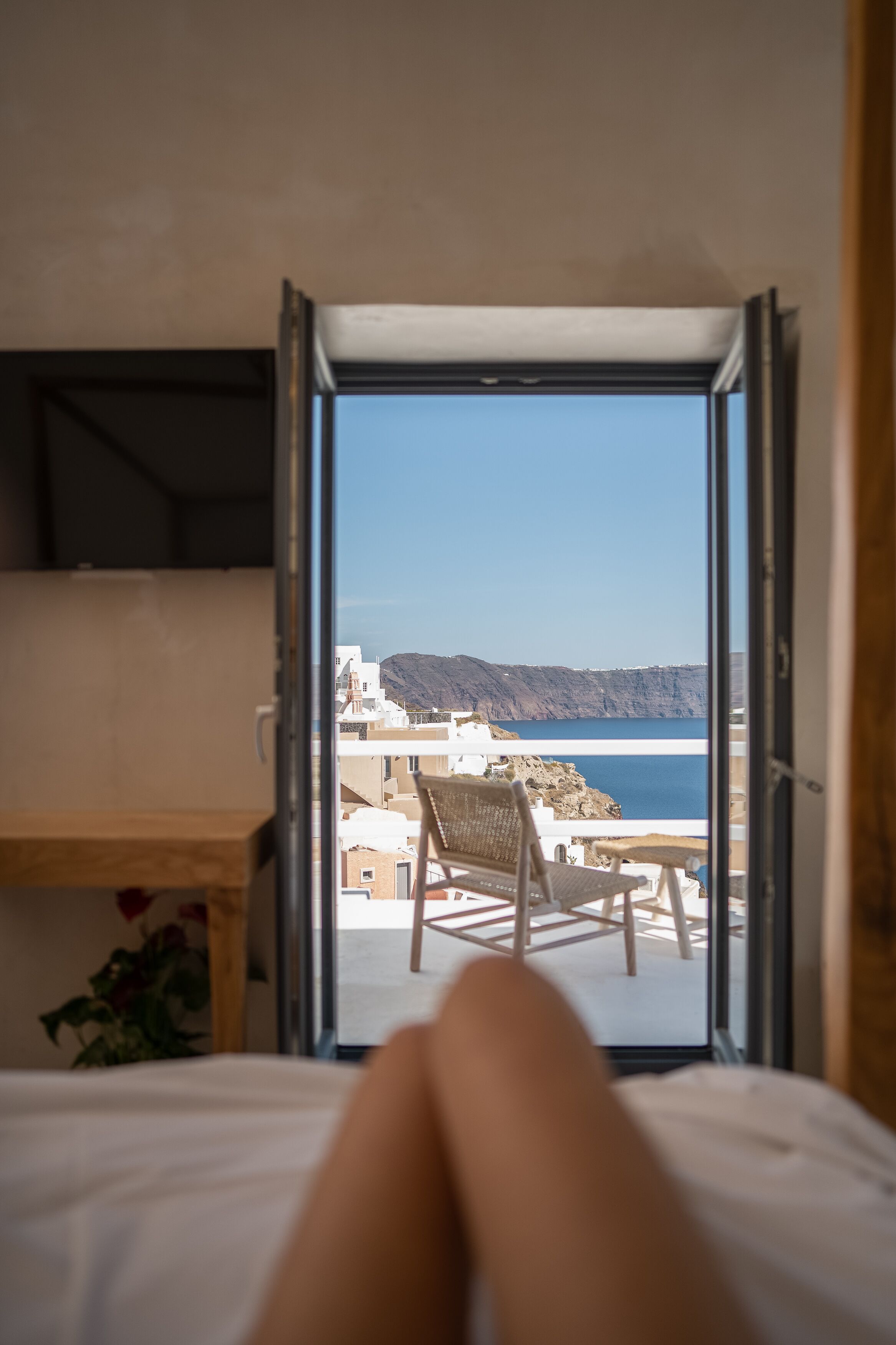 A room with a bed, a wooden desk, and a view of the sea and buildings through an open door.