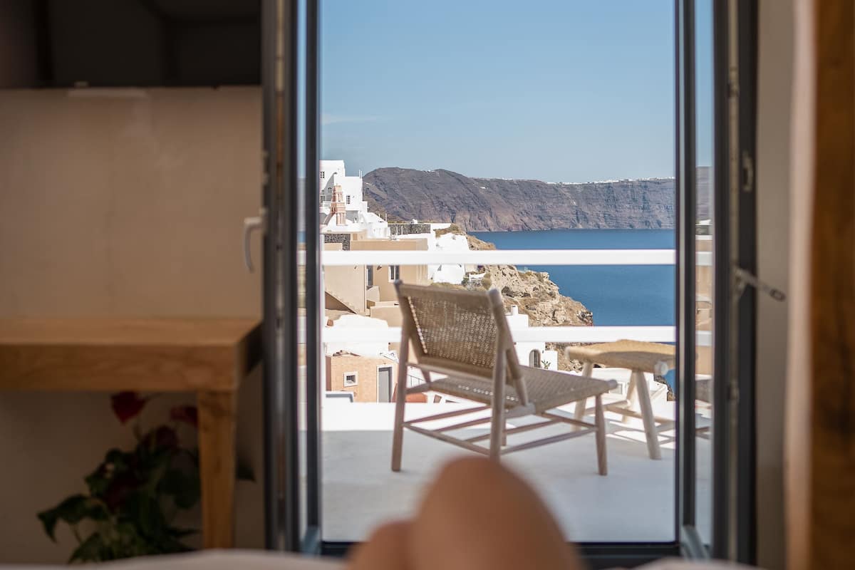 A room with a bed, a wooden desk, and a view of the sea and buildings through an open door.