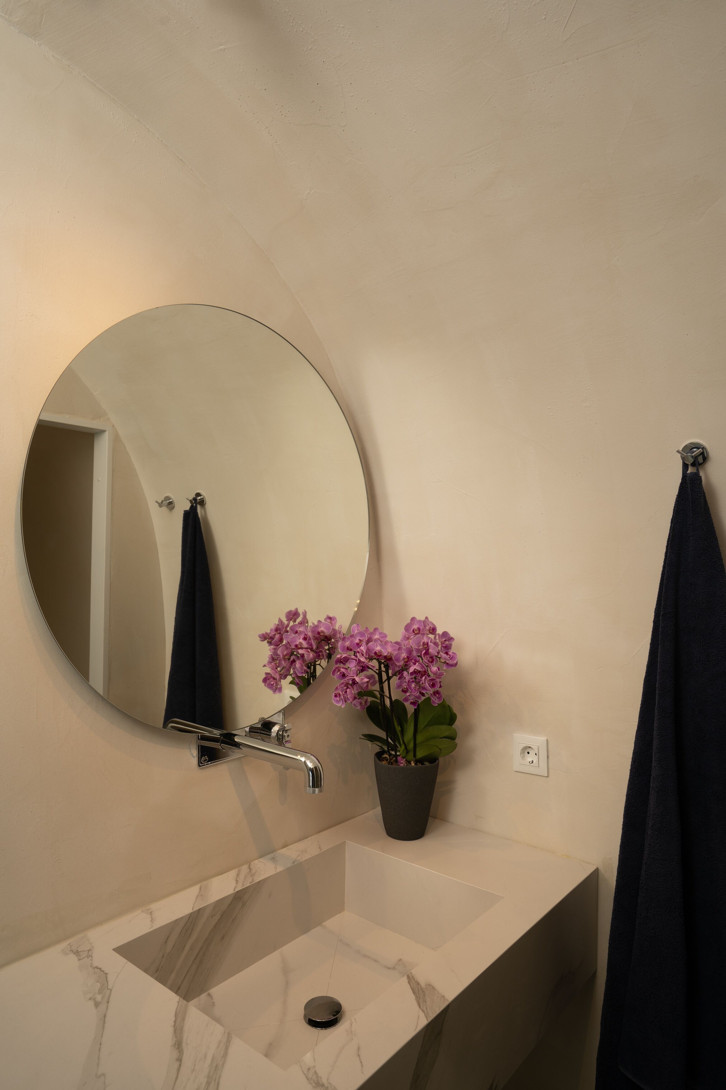 Une salle de bain avec un lavabo en marbre, un miroir rond, une orchidée en pot et un porte-serviettes.