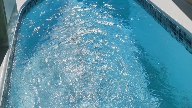Une piscine aux bords incurvés, avec une eau d’un bleu cristallin, entourée de bâtiments blancs, et offrant une vue sur la mer et les montagnes en arrière-plan.