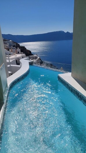 Una piscina a forma di curva con acqua cristallina, circondata da edifici bianchi, e vista sul mare e sulle montagne sullo sfondo.
