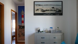 Ein Flur mit einem gerahmten Foto von Strand und Leuchtturm, einer grauen Ablage mit Dekorationsgegenständen und einem blauen Stuhl mit Kissen in Form eines Seesterns.