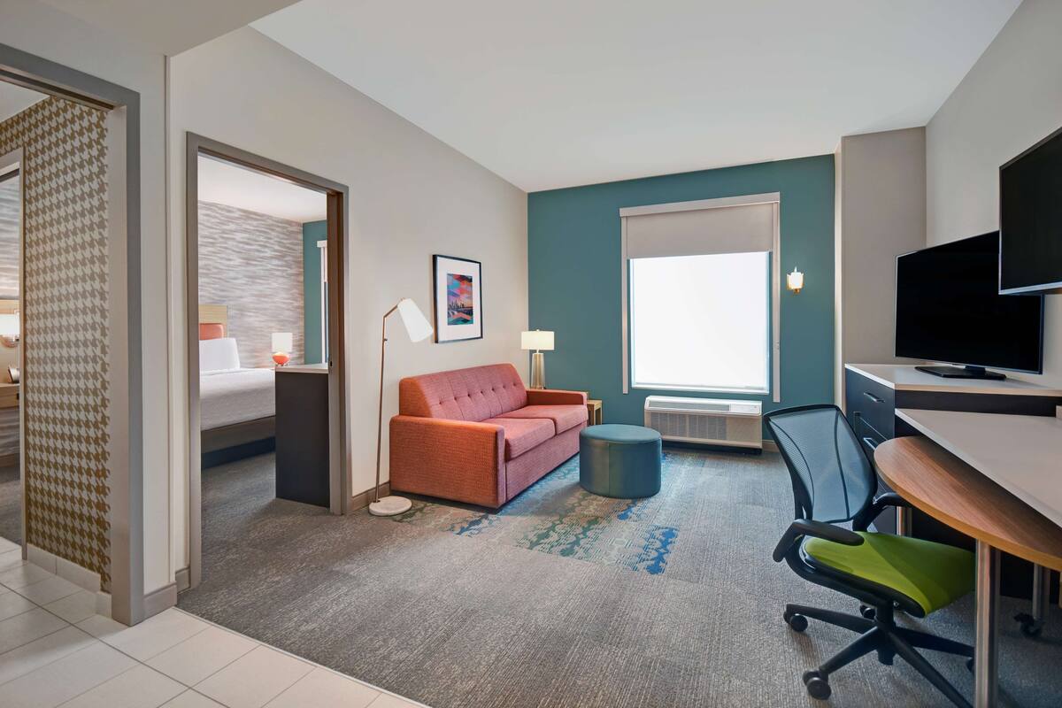 A hotel room with a bed, a red sofa, a desk with a computer, a TV, and a window with a view.