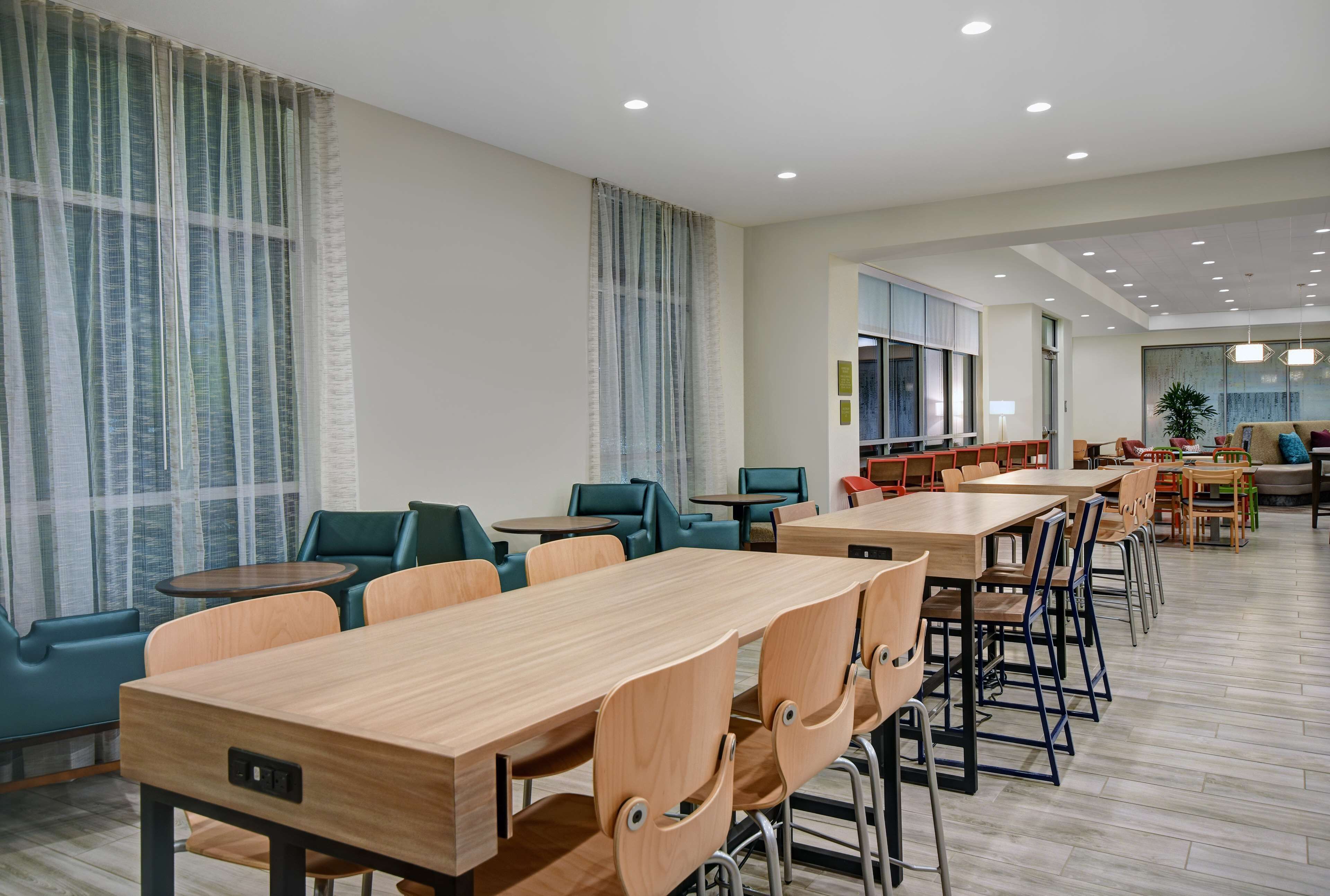 A modern dining area with wooden tables and chairs, large windows with sheer curtains, and a bright, well-lit interior.