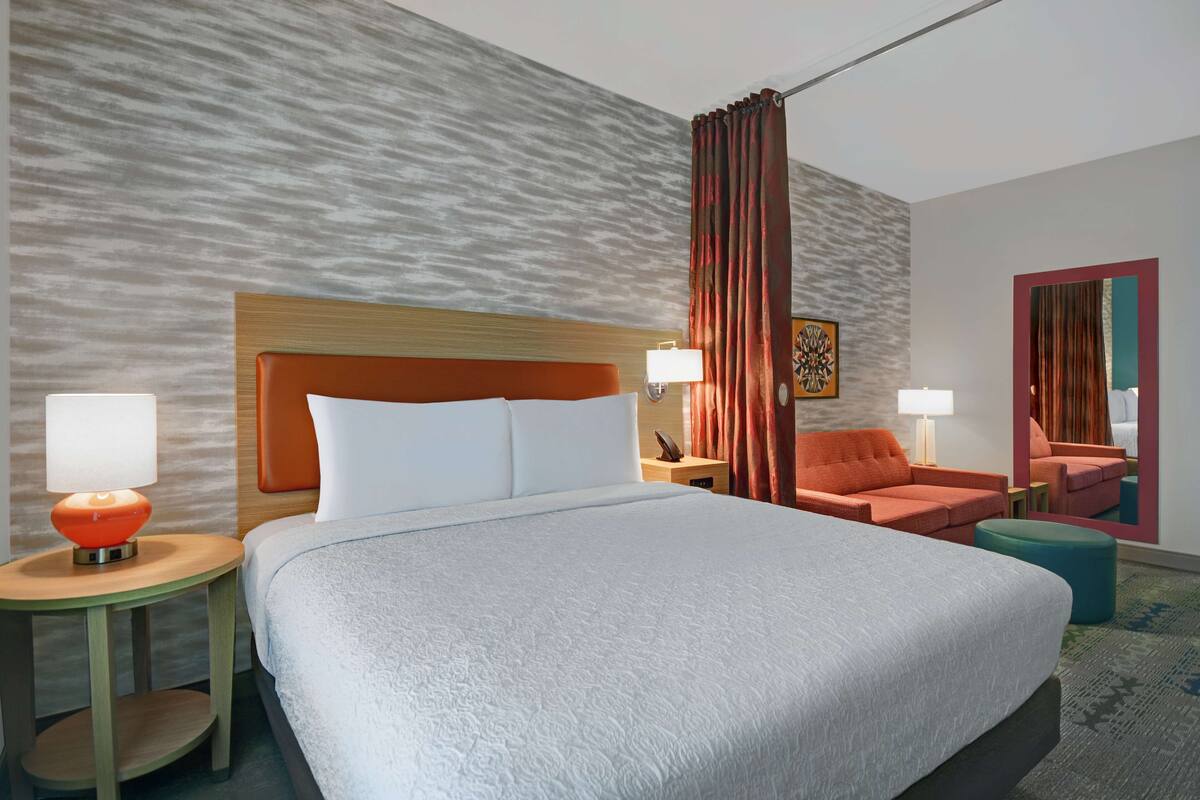 A hotel room with a large bed, two nightstands with lamps, a wooden headboard, a red curtain, a small orange chair, and a mirror on the wall.